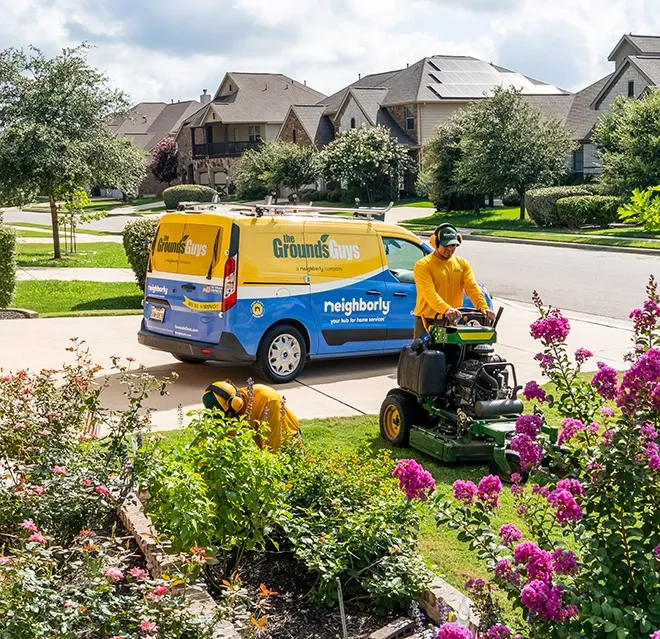 The Grounds Guys service professional using a riding lawn mower to cut the grass of a home's front yard, with a branded van behind him.