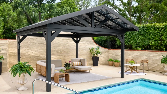 A pergola installed on an inground pool deck, covering outdoor furniture, with hedges and a low stone wall framing the pool area.