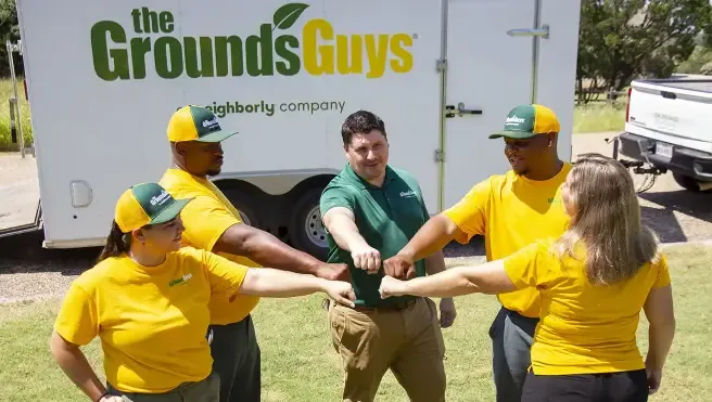 The Grounds Guys crew standing in a circle with their hands in the center, in front of a branded company trailer.